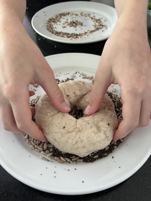 Bagel Fertigung. Man sieht zwei Hände die den Bagel in die Bestreuung drücken.