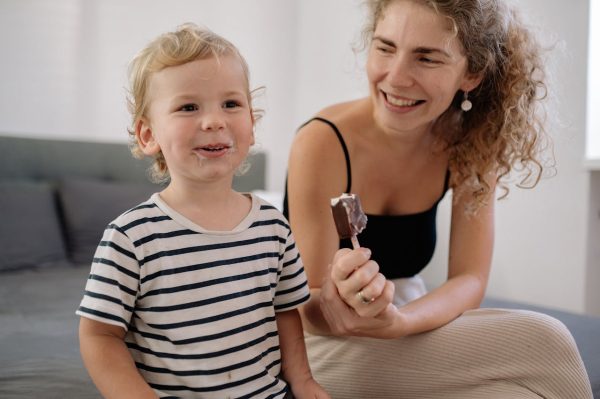 Lachendes Kleinkind mit Eis, im Hintergrund die Mama